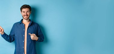 Mutlu gülümseyen adam sol logoyu işaret ediyor, reklamını yapıyor, mavi arka planda duruyor..