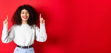Kıvırcık saçlı mutlu genç bir kadın, parmaklarını havaya kaldırıp gülüyor, mükemmel beyaz bir gülümseme sergiliyor, kırmızı arka planda duruyor..