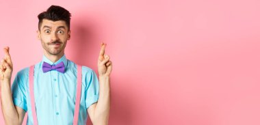 Umut dolu genç bir adam dilek diliyor, parmaklarını çapraz yapıp gülümsüyor, iyimserlikle sonuçları bekliyor, pembe arka planda duruyor..