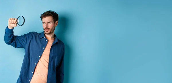 Image of young man searching for something, looking at empty space with magnifying glass, standing on blue background.