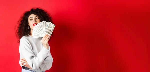 Beautiful rich woman looking sensual aside, waving at herself with dollar bills fan, standing seductive on red background.