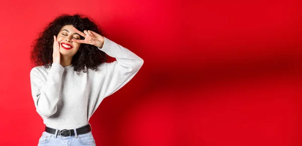 Beauty. Pretty lady with curly hair and red lips, touching face with makeup and showing v-sign on eye, smiling carefree, standing against studio background.