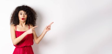 Kırmızı elbiseli ve makyajlı şaşırmış bir kadın parmaklarını doğru gösteriyor, logoyu gösteriyor ve ilgi çekici görünüyor, beyaz arka planda duruyor..