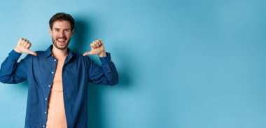 Kendi reklamını yapan mutlu genç adam, kendini işaret ediyor ve gülümsüyor, mavi arka planda profesyonel gibi duruyor..
