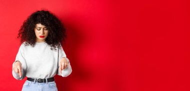 Kıvırcık saçlı endişeli genç bir kadın, yere bakıyor ve endişeli, tereddütlü bir yüzle, kırmızı arka planda duruyor..