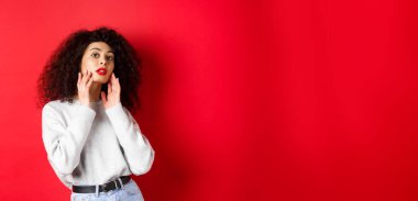 Kırmızı dudaklı güzel ve şık kıvırcık kadın, mükemmel genç yüze dokunuyor ve kameraya şehvetli bakıyor, kırmızı arka planda duruyor..