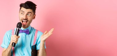 Erkek sunucunun konuşma yaparken görüntüsü, koltuk altında pano olan mikrofon ile konuşması, insanları şenlikli bir olaya dahil etmesi, pembe arka planda dikilmesi..