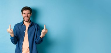 Ignorant and rude guy showing middle finger and smiling, say fuck you, standing on blue background. Copy space