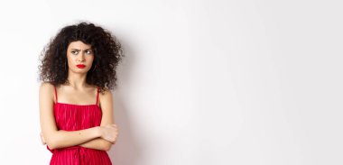 Şüpheli ve gönülsüz zarif bir kadın, kırmızı elbise giyiyor ve kaşlarını çatıyor, inançsızlıkla sol tarafa bakıyor, beyaz arka planda duruyor..