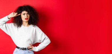 Sen aptal mısın? Kıvırcık saçlı, kızgın ve kafası karışık bir kız. Kafasını işaret ediyor ve azarlıyor. Deli ya da tuhaf, kırmızı arka planda duruyor..