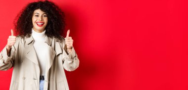 Mutlu görünen genç bir kadının portresi, trençkot giyiyor ve başparmak kaldırıyor, evet diyor, onaylıyor ve övüyor güzel, kırmızı bir arka plan.