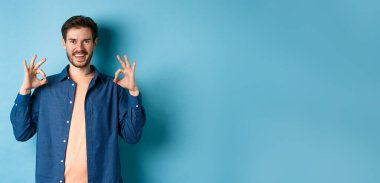 Tatmin olmuş genç adam gülümsüyor ve iyi bir jest yapıyor, iyi bir şeyi onaylıyor, mükemmel bir şeyi övüyor, mavi arka planda duruyor..