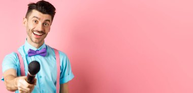 Bıyıklı ve papyonlu gülümseyen adam yorum ya da röportaj istiyor mikrofonla elini uzatıp pembe arka planda duruyor..