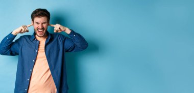 Sinirli genç adam can sıkıcı sesleri engellemek ve kameraya bağırmak için kulaklarını kapatmış. Kaşlarını çatmış ve öfkeli görünüyor. Mavi arka planda duruyor..
