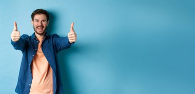 Yakışıklı, gülümseyen ve onay veren genç adam, mutlu bir yüzle evet de, mavi arka planda dur..