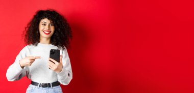 Akıllı telefonlu neşeli bir bayan. Telefonunu işaret ediyor. Tatmin olmuş bir şekilde gülümsüyor..