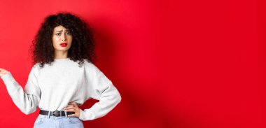 Kafası karışmış ve şüpheci bir kadın, kıvırcık saçlı, kaşlarını çatan ve el kaldıran, kırmızı arka planda duran garip bir şeyi anlayamıyor..