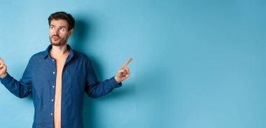 Kararsız bir adam günlük kıyafetlerini giyiyor, seçimler yapıyor, parmaklarını yana çeviriyor ve dalgın bakıyor, mavi arka planda duruyor..