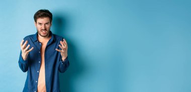 Annoyed and angry man raising hands and clench teeth outraged, stare pissed-off at camera, going to kill someone, standing on blue background.