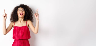 Heyecanlı beyaz kadın kıvırcık saçlı, kırmızı dudaklı, şaşkın bir gülümsemeyle bakıyor ve işaret ediyor, tanıtım teklifini kontrol ediyor, beyaz arka planı var..