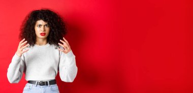 Kızgın beyaz kadın, kaşlarını çatıyor ve ellerini kaldırıyor. Kırmızı arka planda dikilen sinir bozucu birini boğmak ya da öldürmek istiyor..