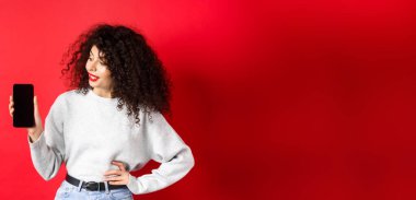 Şık, kıvırcık saçlı, boş akıllı telefon ekranı gösteren, alışveriş uygulamasını gösteren, kırmızı arka planda duran genç bir kadının portresi..
