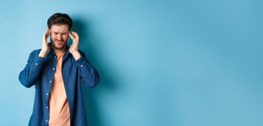 Hasta hisseden, başıyla temas eden, baş ağrısı çeken, baş dönmesi hisseden, mavi arka planda duran bir adam..