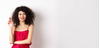 Güzellik ve moda. Kıvırcık saçlı, makyajlı, kırmızı elbiseli, el sallayan, selam veren, beyaz arka planda duran gülümseyen kadın..