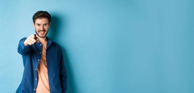 Kendinden emin gülümseyen adam seni seçiyor ya da davet ediyor, kamerayı işaret ediyor kararlı, mavi arka planda duruyor..