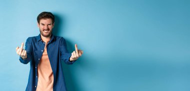 Angry caucasian man say fuck you and showing middle fingers, make rude gesture with pissed-off face, standing annoyed on blue background.