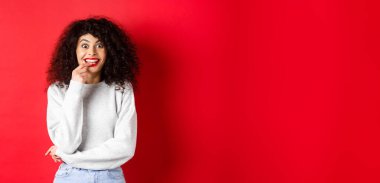 Heyecanlı ve entrikacı bir kadın bir şeyler denemeye hevesli, baştan çıkarıcı bakışlar ve tırnak yemek eğlenceli, kırmızı arka planda durmak.