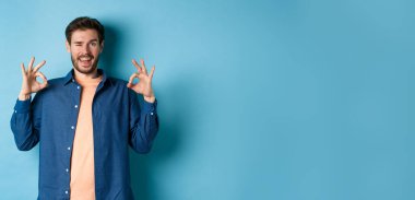 Gülümseyen ve göz kırpan mutlu adam Tamam işareti, evet de, onayla ve kabul et mavi arka planda duran iyi bir şeyi.