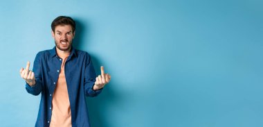 Angry rude guy showing middle finger and saying fuck you, swearing and staring furious at camera, standing on blue background.