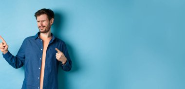 Şüpheci genç adam hayal kırıklığıyla sol tarafı işaret ediyor, kötü bir şeyden korkup mavi arka planda üzgün duruyor..