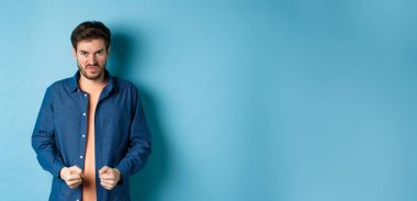Angry and pissed-off man clench fists and stare with disdain at camera, trying to keep himself together, look with rage, standing on blue background.