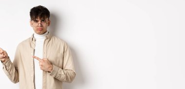 Üzgün ve huysuz çocuk, solu işaret ediyor, çocuksu bir ifadeyle somurtuyor, kötü bir şeyden şikayet ediyor, beyaz arka planda duruyor..