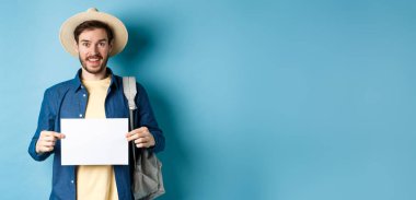 Yazlık şapkalı neşeli turist, boş bir kağıt parçası gösteriyor ve gülümsüyor, sırt çantasıyla otostop çekiyor, mavi arka planda duruyor..