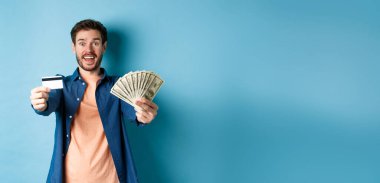 Dolarlarla ve kredi kartıyla elini uzatan neşeli beyaz adam, kameraya mutlu bir şekilde gülümsüyor, mavi arka planda duruyor..