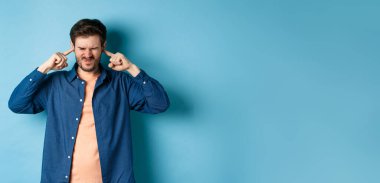 Sinirli genç adam sesi, parmaklarıyla kulaklarını kapatıyor ve yüzünü buruşturuyor, yüksek sesli müzikten rahatsız oluyor, mavi arka planda duruyor..
