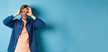 Komik beyaz adam el dürbününden bakıp gülümsüyor, ilginç bir şeye bakıyor, mavi arka planda şaşkın şaşkın duruyor..