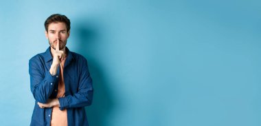 Modern adam kameraya şşş yapıyor, parmaklarını dudaklarına bastırıyor ve bir sır söylüyor, tabu işareti yapıyor, mavi arka planda duruyor..