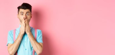 Şok olmuş bir adam bakıyor, elleriyle ağzını kapatıyor ve pembe arka planda durup kameraya bakıyor..