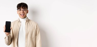 Teknoloji ve online alışveriş konsepti. Gözlüklü neşeli yakışıklı adam boş akıllı telefon ekranı gösteriyor ve gülümsüyor, uygulamayı gösteriyor, beyaz arka planda duruyor..