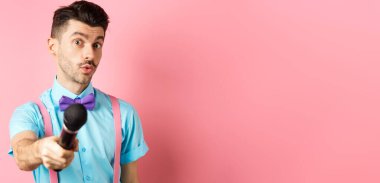 Yakışıklı erkek sunucu mikrofonla elini uzatıyor, röportaj ya da yorum istiyor, pembe arka planda duruyor..