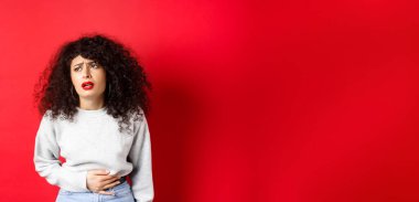 Kadın hasta, eğiliyor ve karnına dokunuyor, karın ağrısı ya da regl sancısı çekiyor, kırmızı arka planda duruyor..