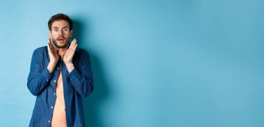 Şok olmuş ve korkmuş genç adam sersemlemiş, ellerini kaldırıyor ve nefes nefese, mavi arka planda duruyor..