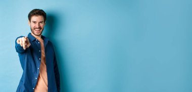 Kendinden emin gülümseyen adam seni seçiyor ya da davet ediyor, kameraya işaret ediyor kararlı, mavi arka planda duruyor. Boşluğu kopyala