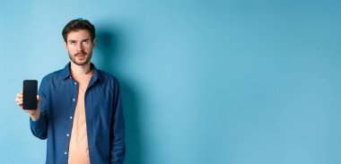 Sinirli genç adam boş akıllı telefon ekranı gösteriyor ve gözleri yuvarlıyor. Mavi arka planda duruyor..