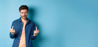 Ignorant and unbothered man showing middle-fingers and looking reluctant at camera, give zero fucks, standing on blue background.
