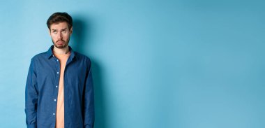 Üzgün adam mızmızlanıyor ve somurtuyor, üzgün bir suratla yukarı bakıyor, mavi arka planda duruyor..